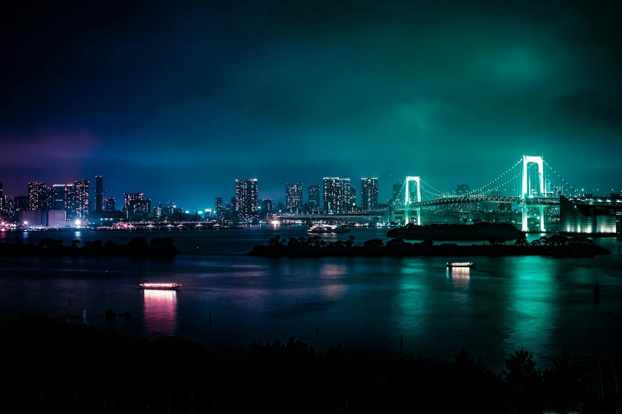 Illuminated Tokyo skyline featuring the iconic Rainbow Bridge at night, reflected in calm bay waters.