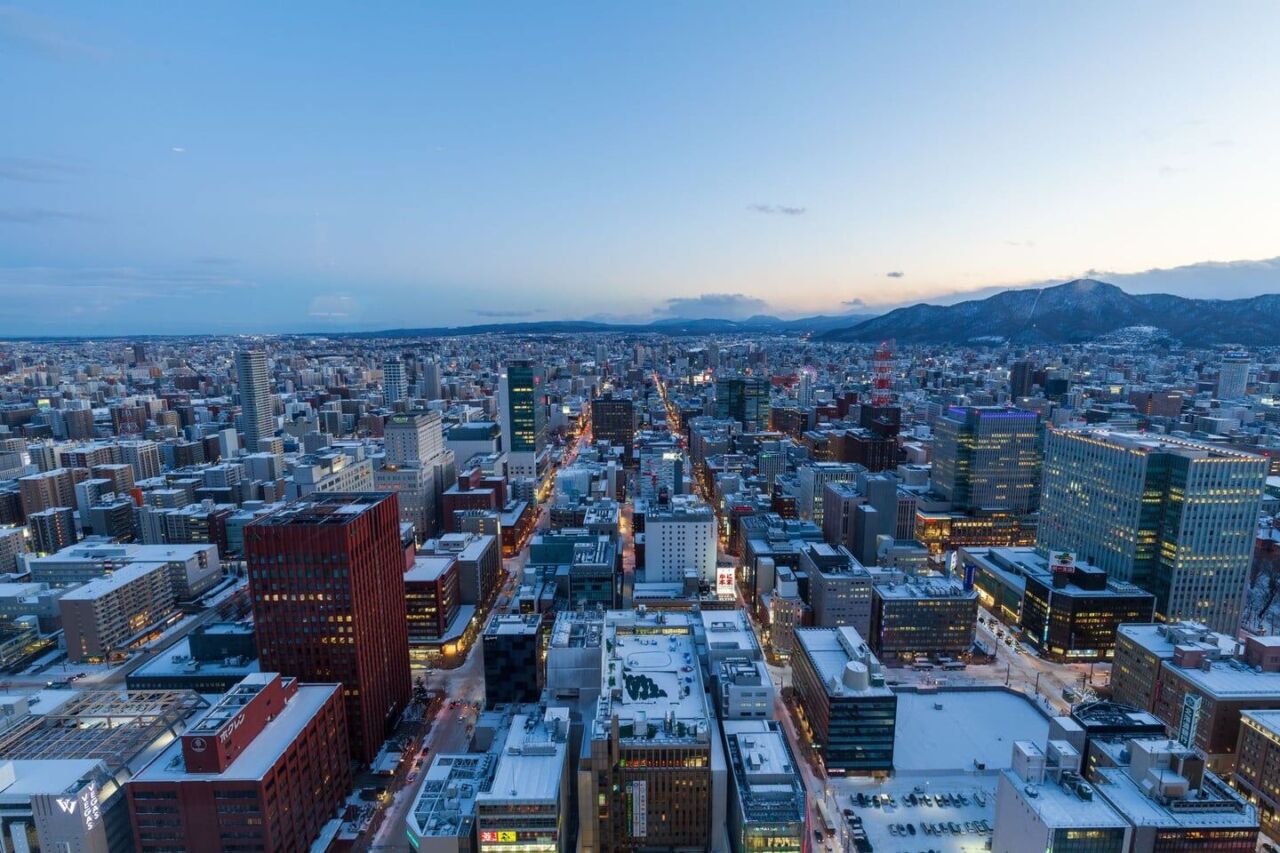 Captivating aerial view of Sapporo, Japan in winter at dusk with snow-covered buildings and distant mountains.