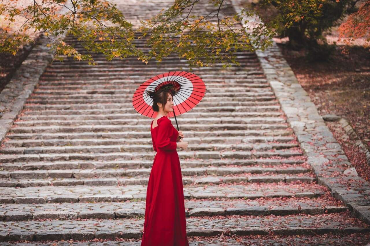 An Asian woman in a red dress holding a traditional umbrella on autumn stone steps.