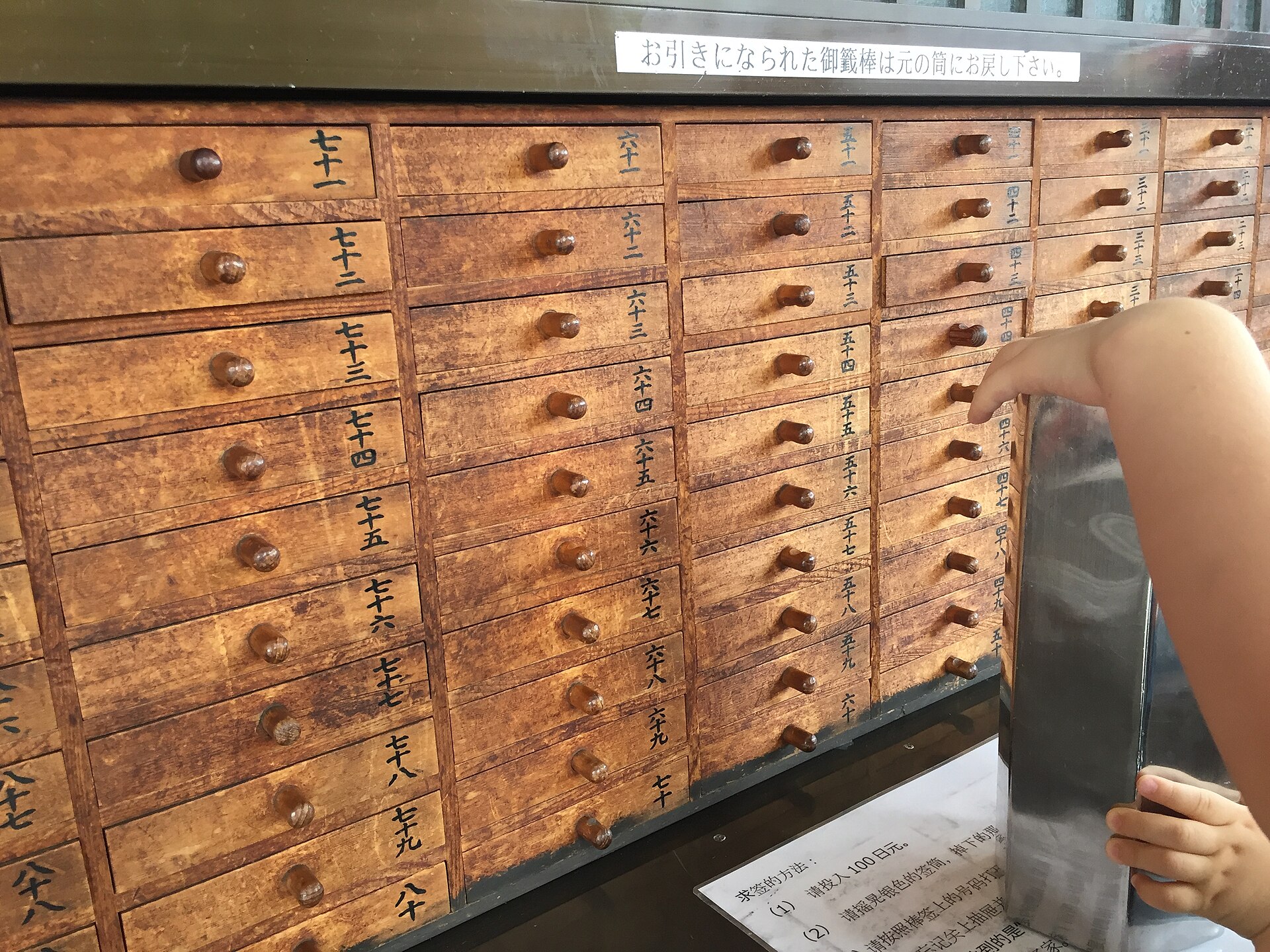 A hand reaching toward numbered wooden drawers of a fortune-slip cabinet, with a metal stand in front