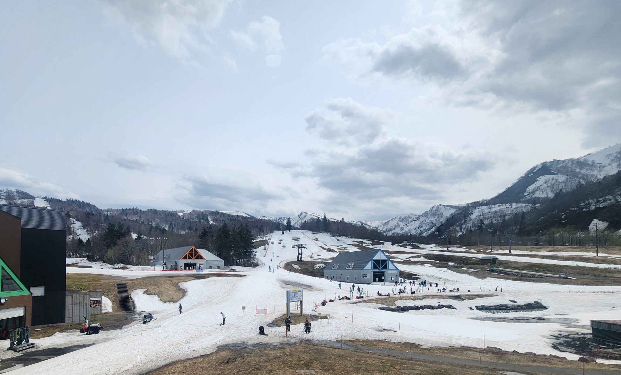 눈이 듬성듬성 남은 스키장. 작은 목조 건물들과 곳곳의 사람들, 뒤로 산과 흐린 하늘.