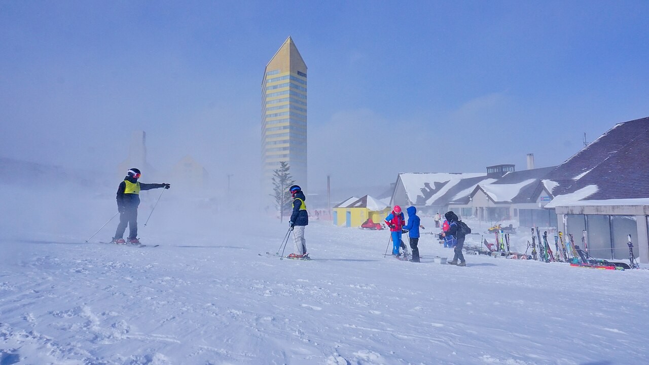 雪原で滑走する黄色いベストの指導者と色鮮やかなウェアの受講者、背景に高い塔と雪屋根の建物