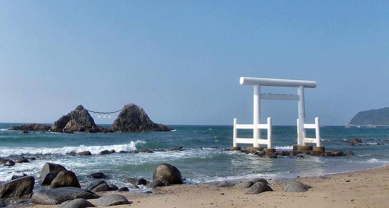 모래사장 끝 바다에 흰 도리이가 서 있고, 금줄로 이어진 두 바위가 보이는 풍경