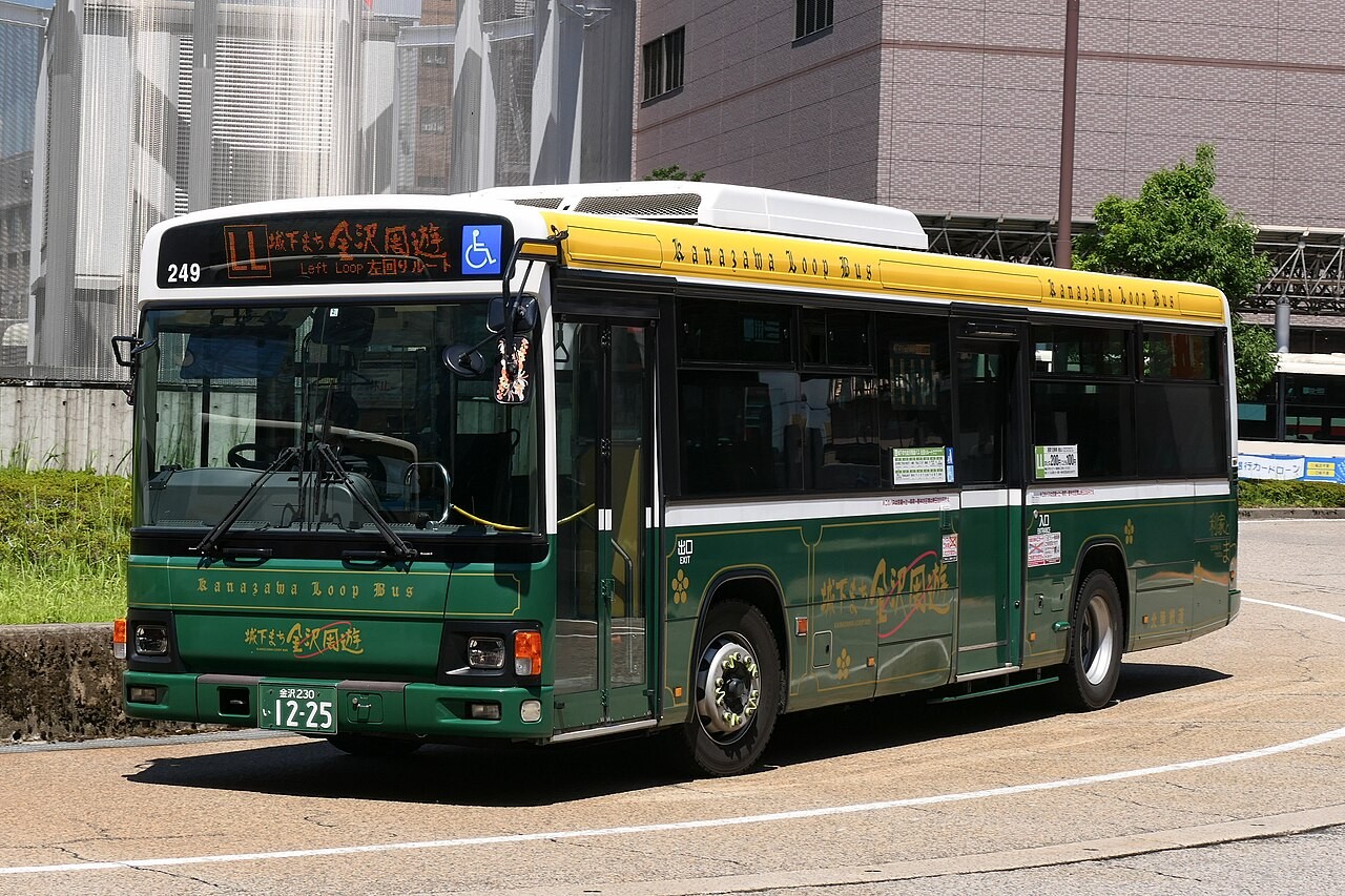 緑と黄色の金沢周遊バスが舗装道路に停車している。背景に建物と植え込み