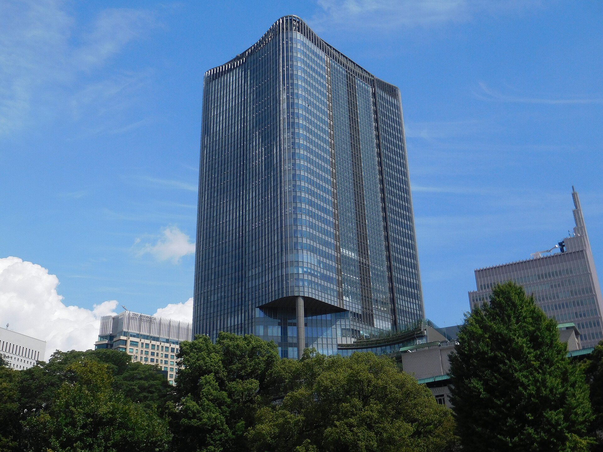 Tall blue glass skyscraper with a pointed top, surrounded by green trees under a blue sky