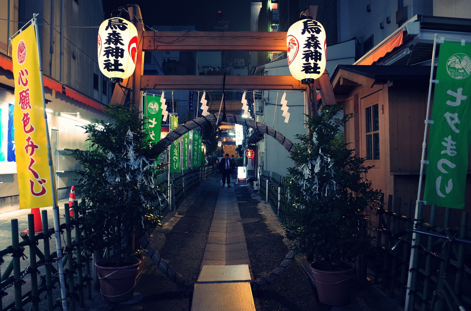 Narrow nighttime shrine approach lined with a torii gate, white lanterns, and green and yellow banners