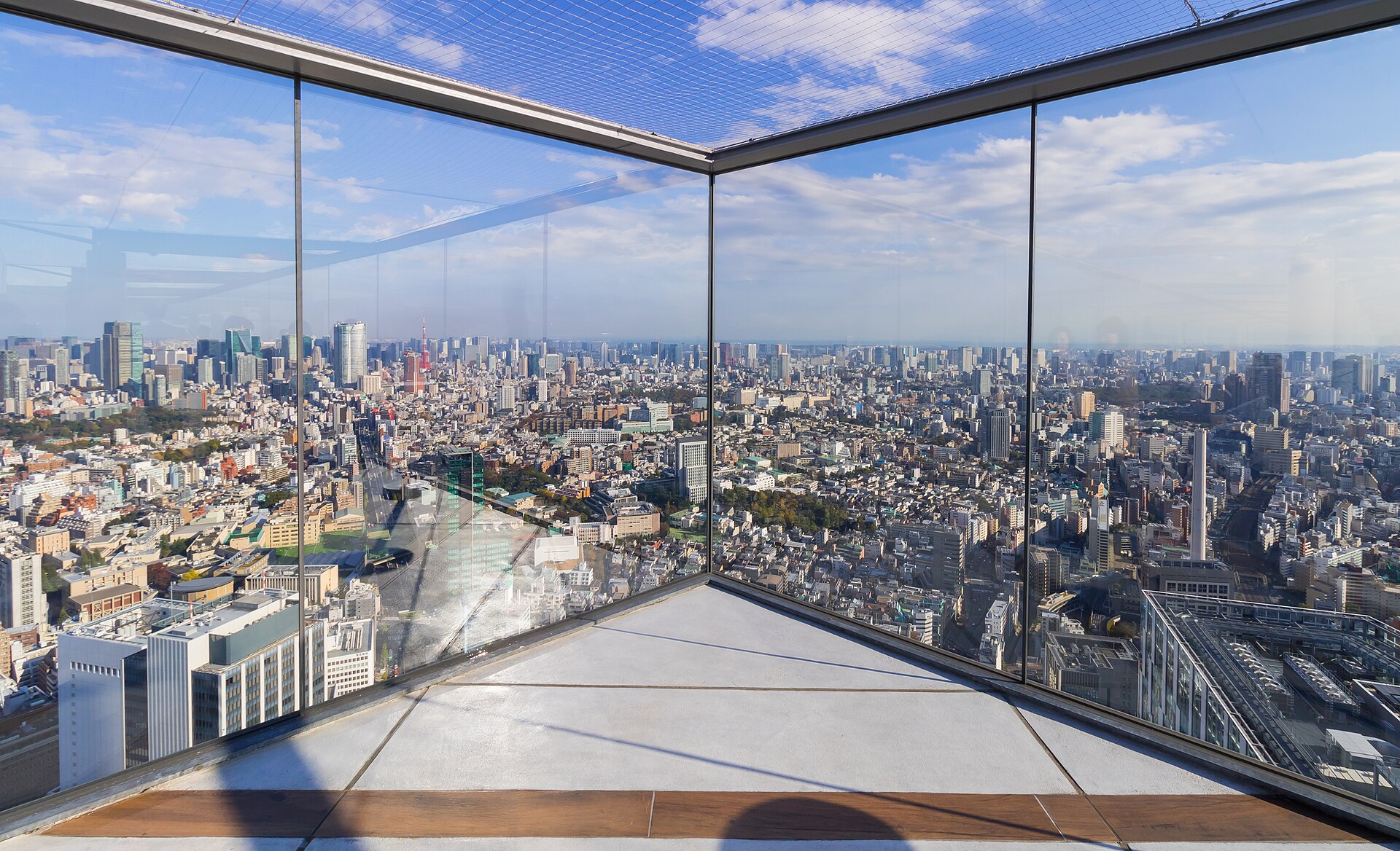 Glass-enclosed observation deck overlooking Tokyo's dense cityscape of high-rises under a blue, cloudy sky.