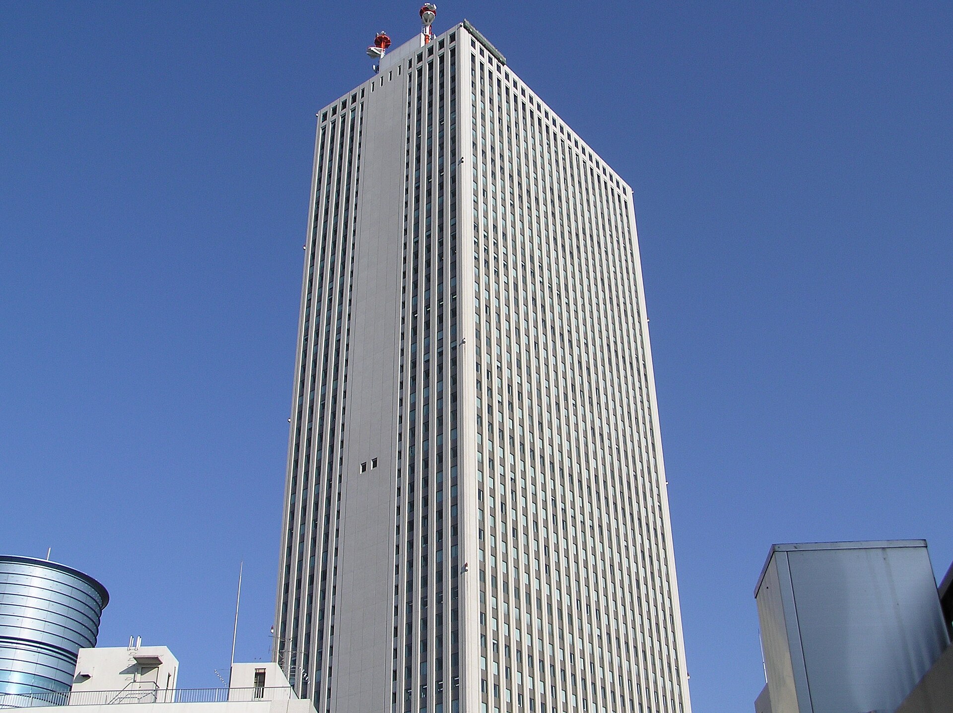 Tall gray-white office tower with vertical window bands under a clear blue sky, with red-and-white antennas on its roof.