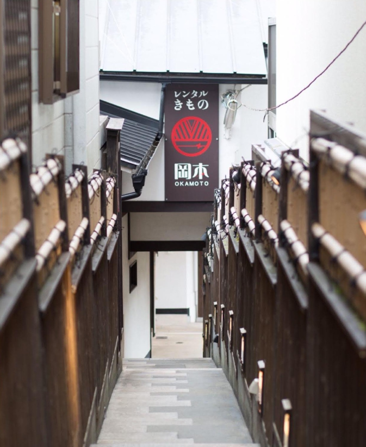 Narrow stone staircase between wooden railings, with a hanging red-and-white Okamoto kimono rental sign ahead.