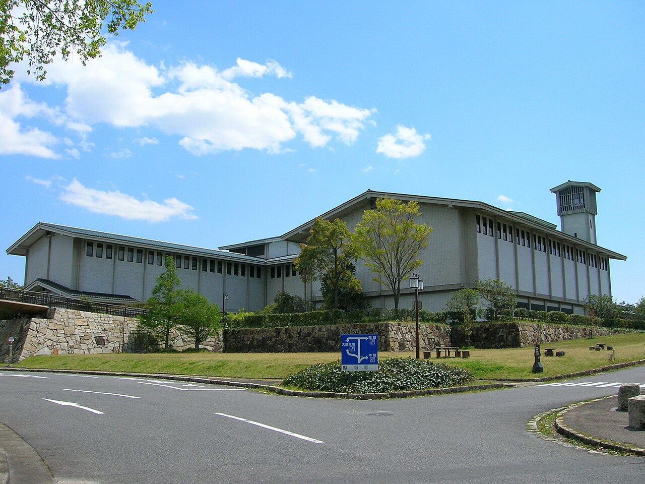 Gray modern museum building with a tower, grass lawn, blue signboard, and road under a clear blue sky