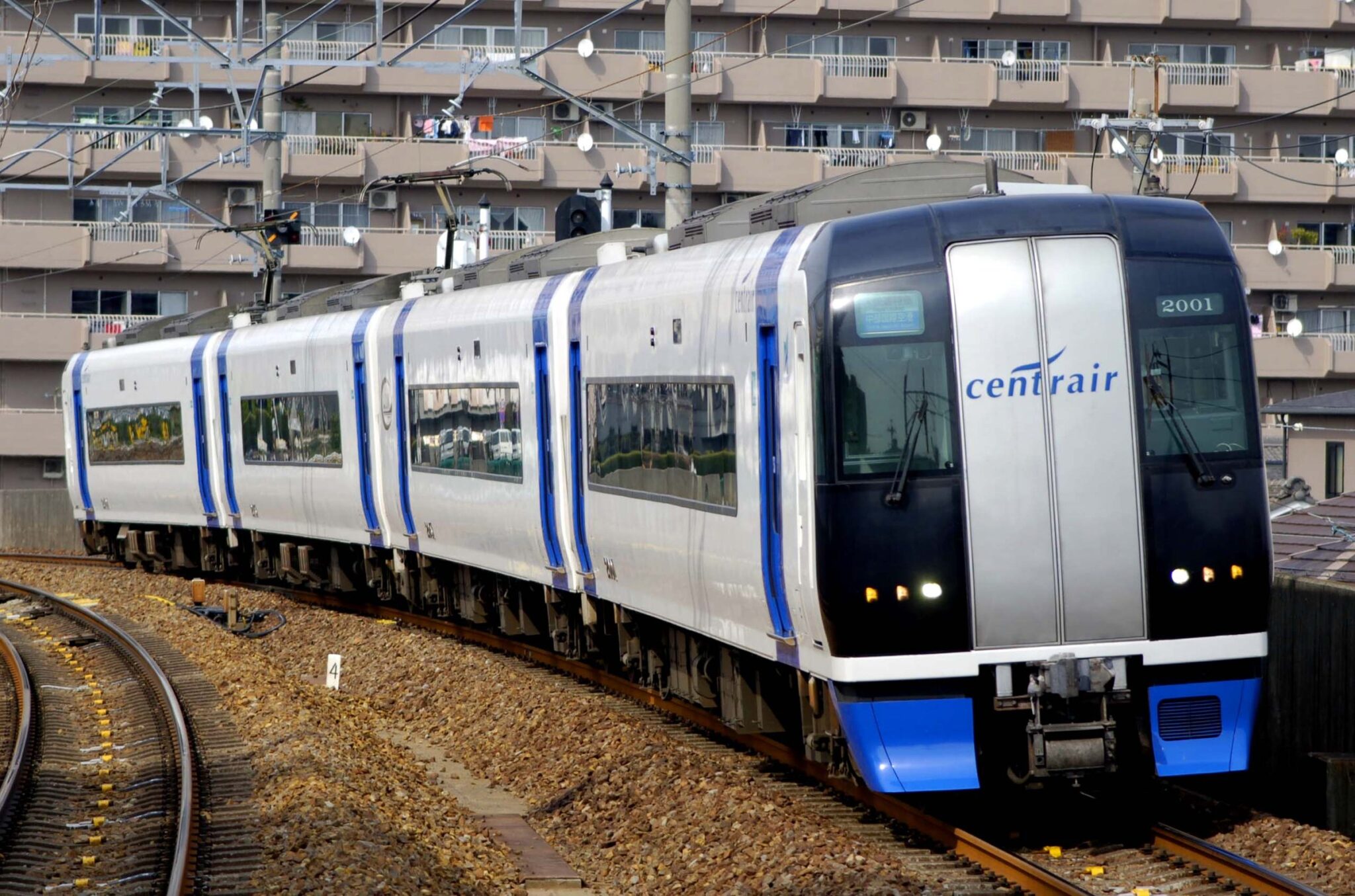 A silver and blue Meitetsu train with 'centrair' branding on tracks, apartment buildings behind