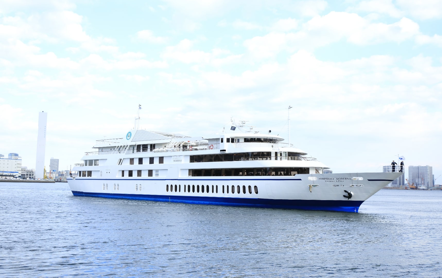 White and blue cruise ship on calm bay water, city buildings and cloudy sky behind