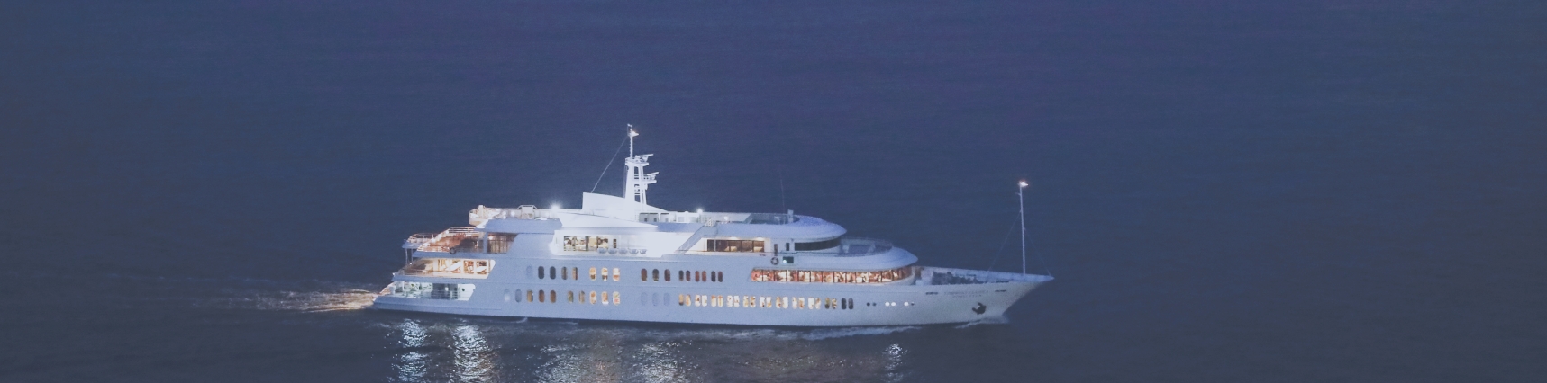A white cruise yacht with illuminated windows sailing across dark blue water at night.
