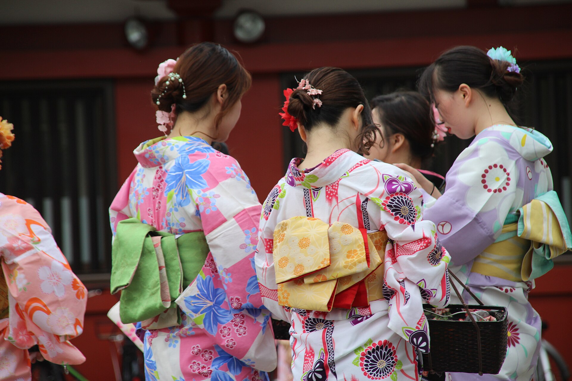 Four women in colorful floral kimonos stand with their backs to the camera before a red building.