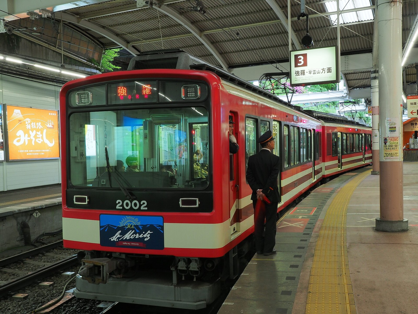 Red-and-cream Hakone Tozan Train stopped at a platform, with a uniformed station attendant standing beside it.