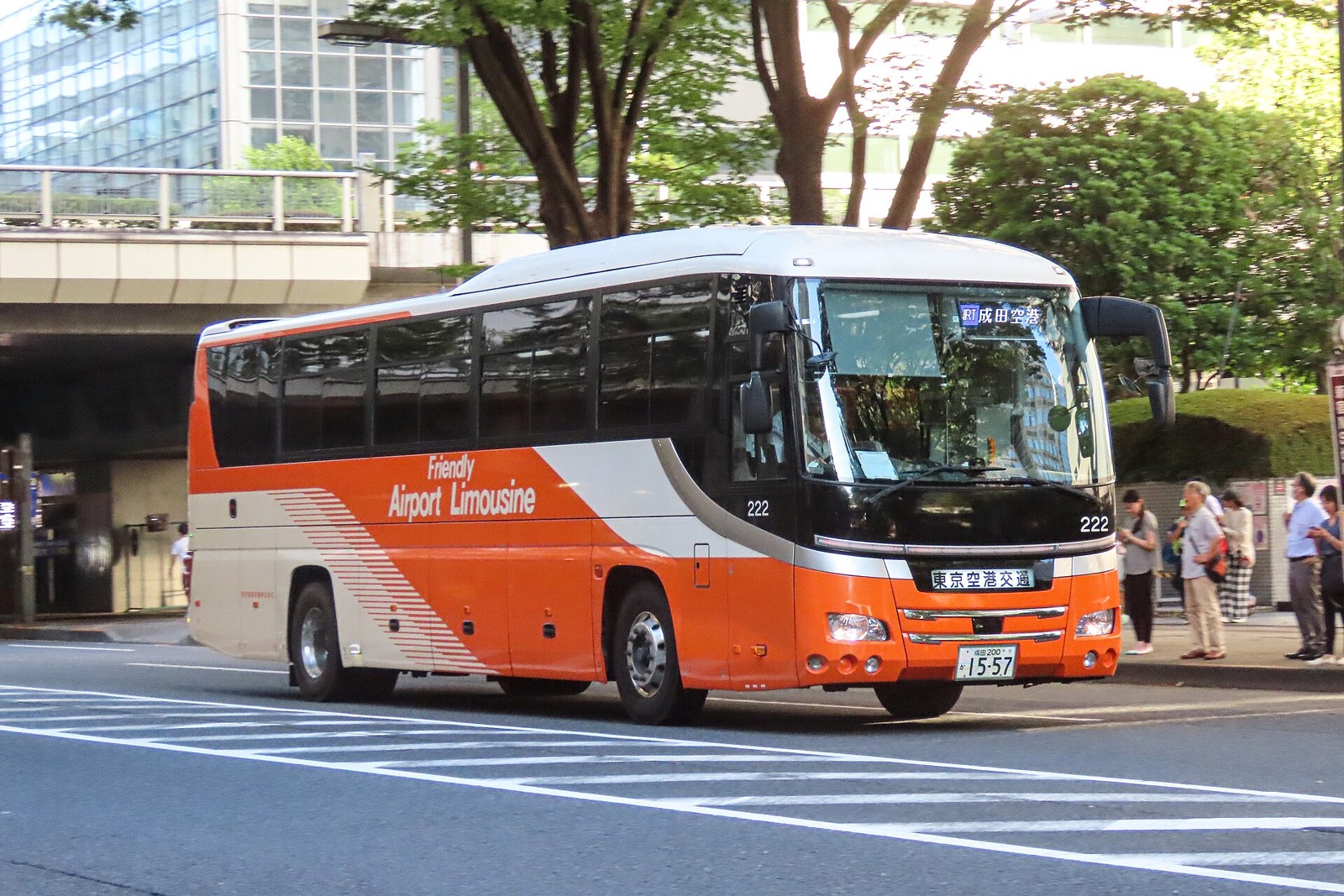 An orange and white Friendly Airport Limousine bus driving on a city street with pedestrians waiting nearby