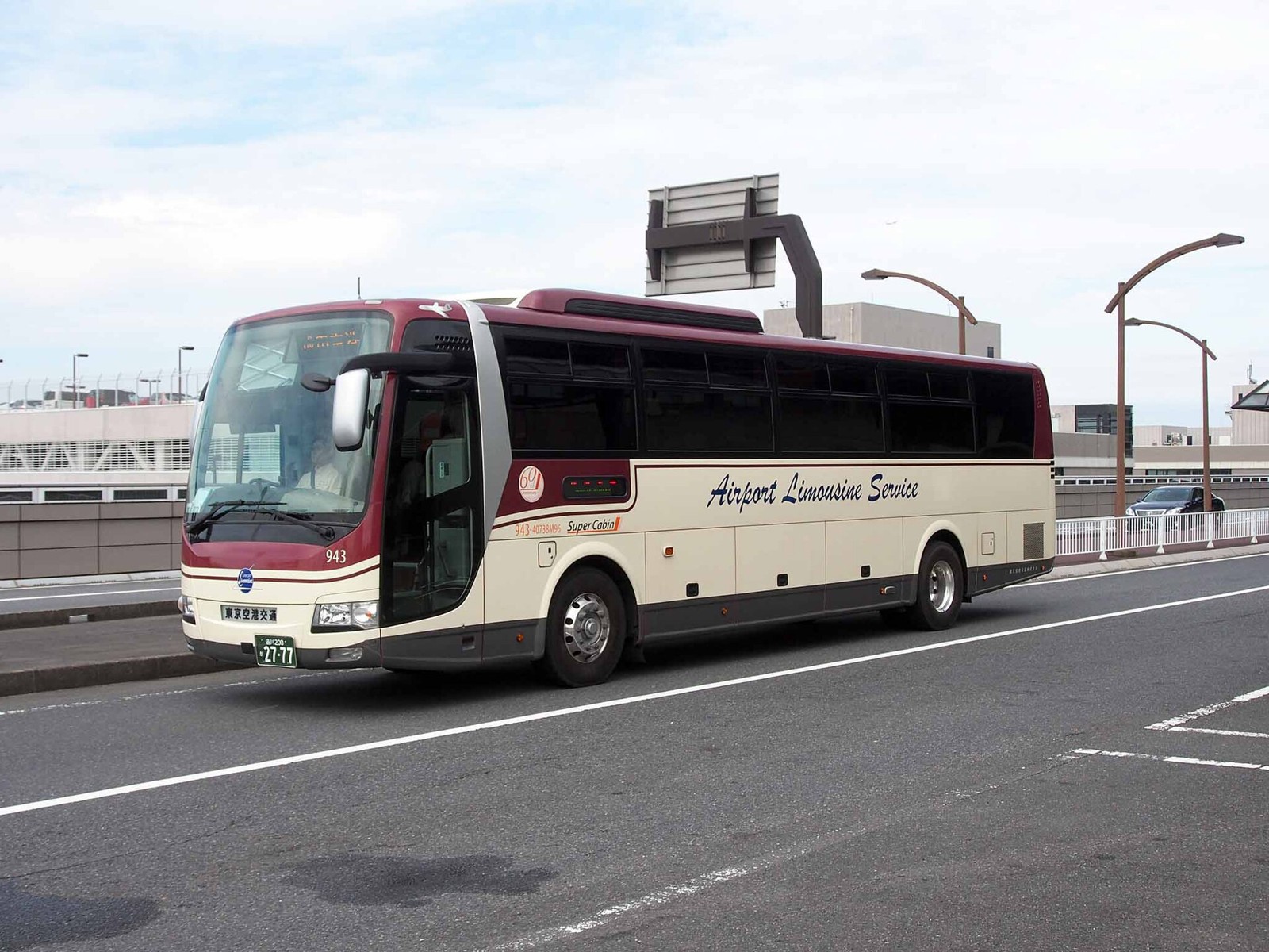 A cream and maroon Airport Limousine Service bus driving on a wide road, with a sign overhead and a building behind.