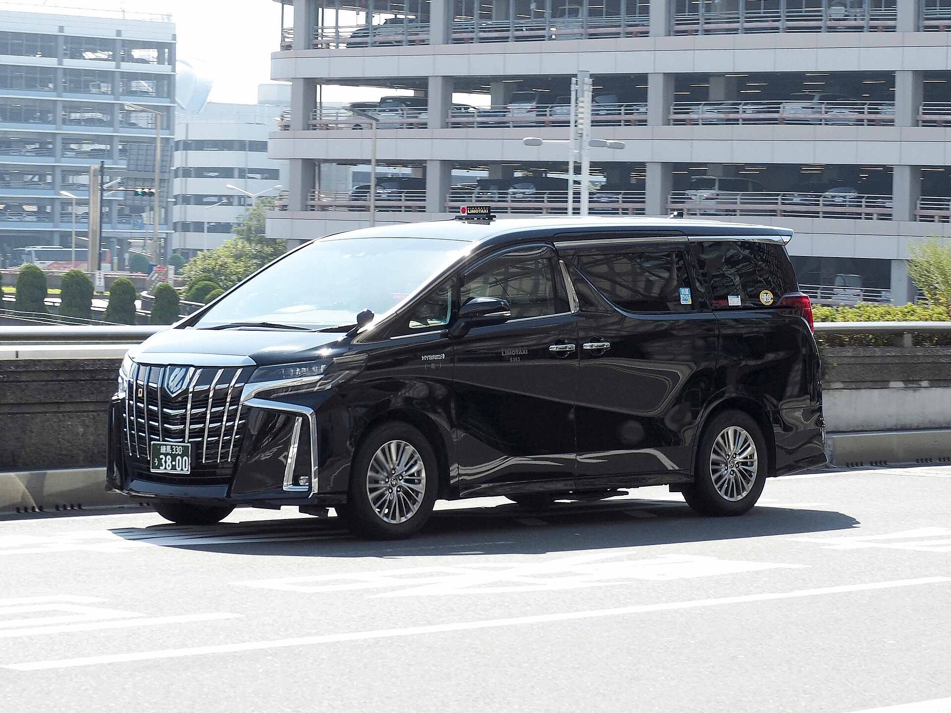 A black Toyota Alphard minivan parked on a road, with a multi-level parking structure behind it.