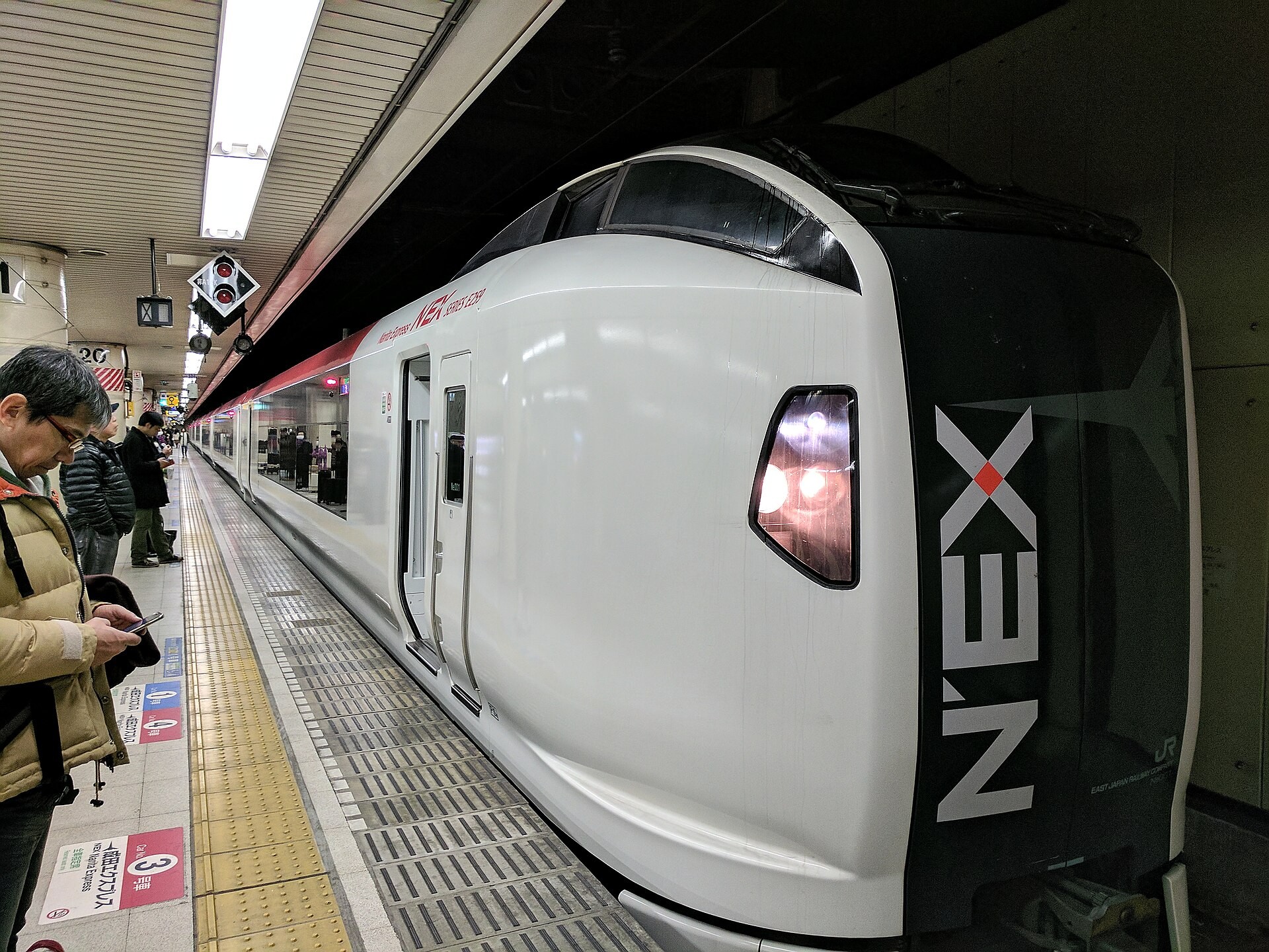 A white N'EX Narita Express train with black front and NEX lettering, stopped at a platform
