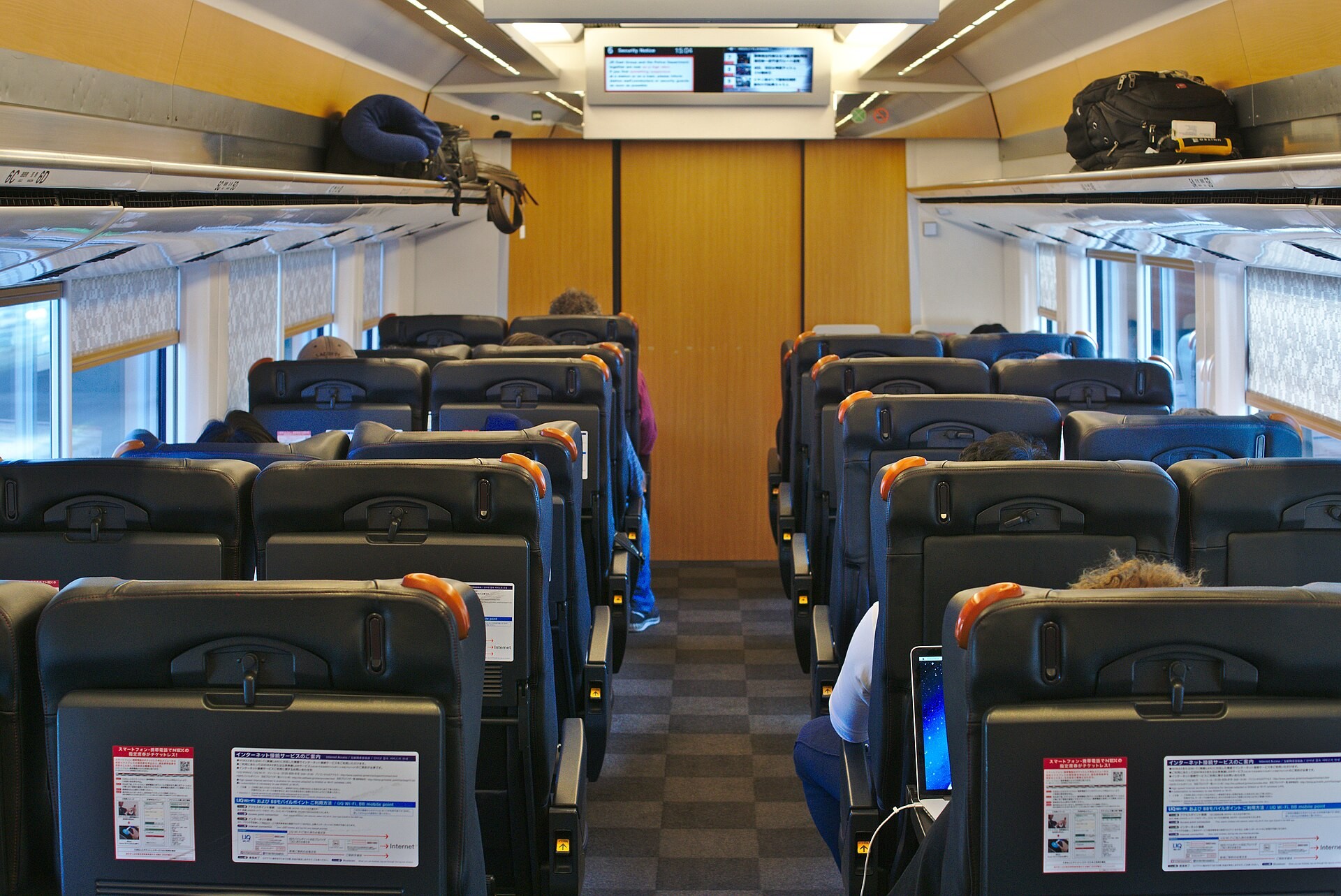 Interior of a train carriage with rows of dark reclining seats, checkered grey carpet, and overhead luggage racks
