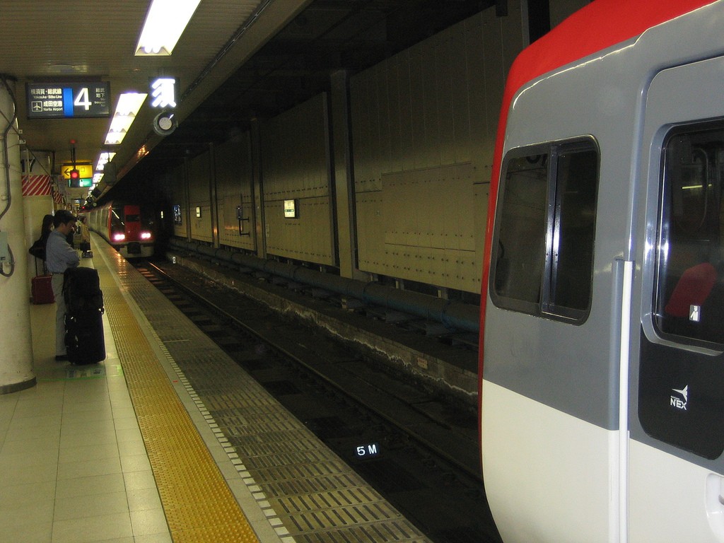 Platform at Narita Airport station with a red-and-white NEX train on the right and a man waiting beside rolling luggage.