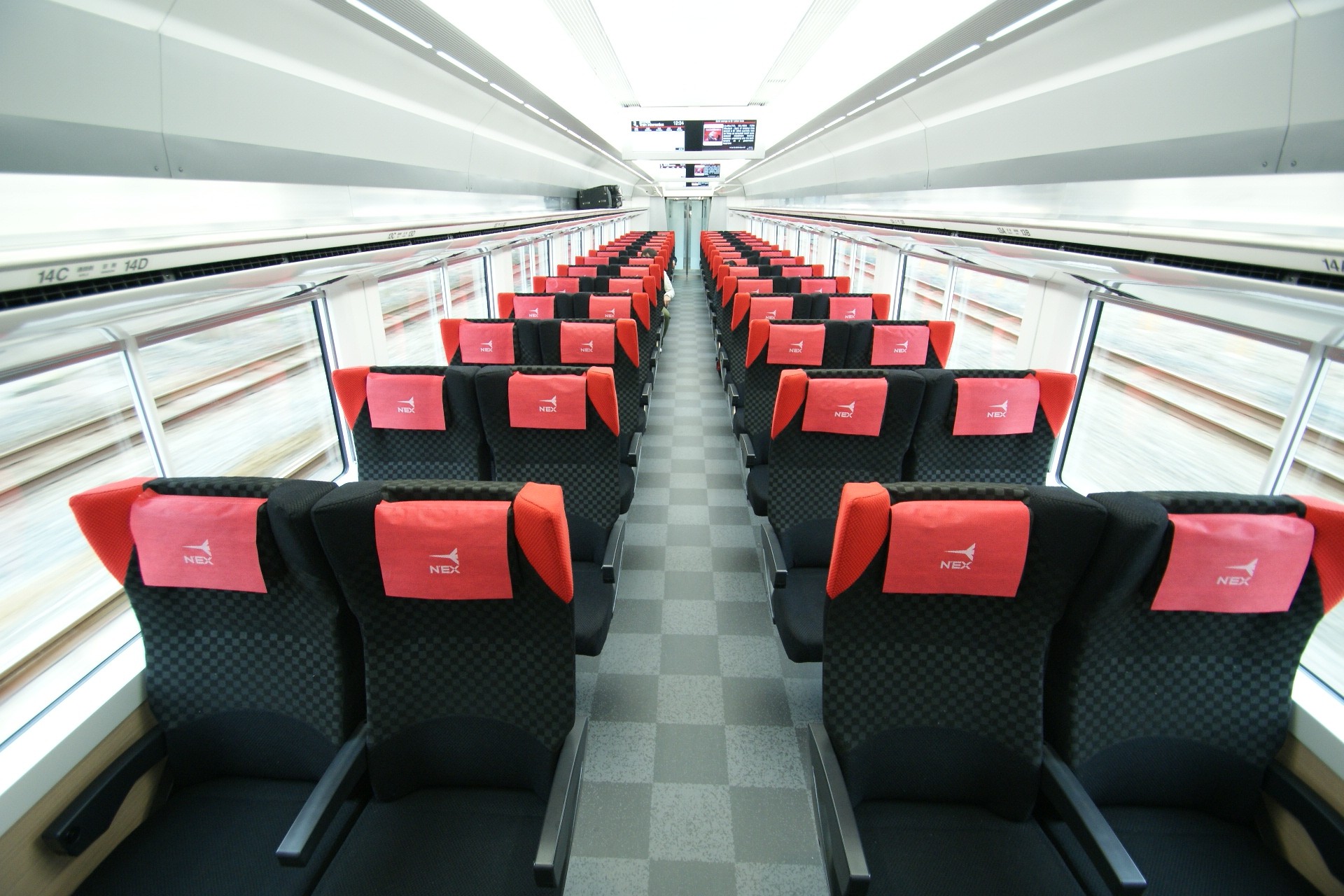Interior of an N'EX train car with rows of black checkered seats and red headrest covers