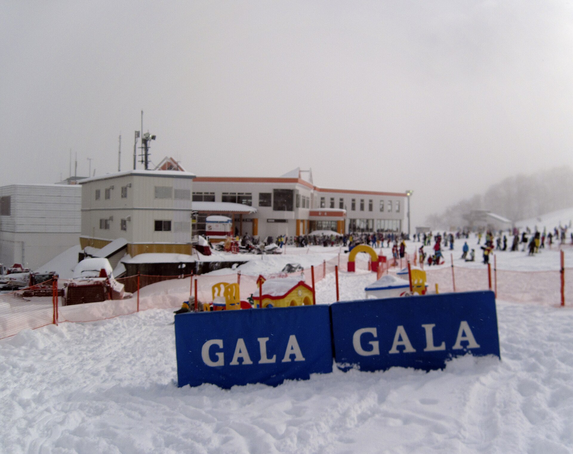 Blue GALA signs on snow in front of a white building with skiers and colorful playground equipment