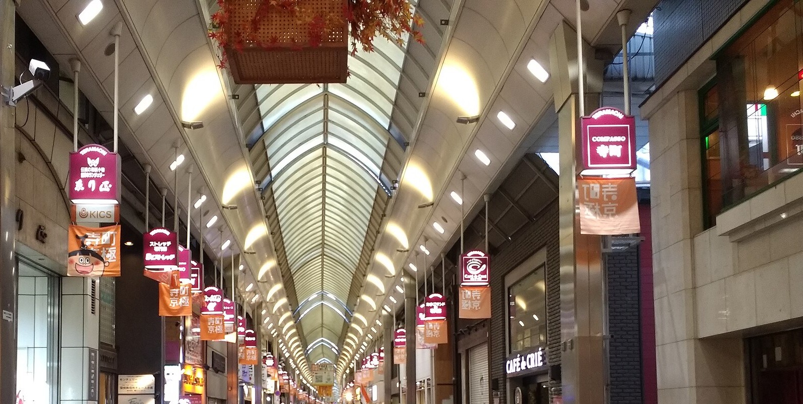 Covered shopping arcade interior with arched glass ceiling and rows of hanging red and orange signs