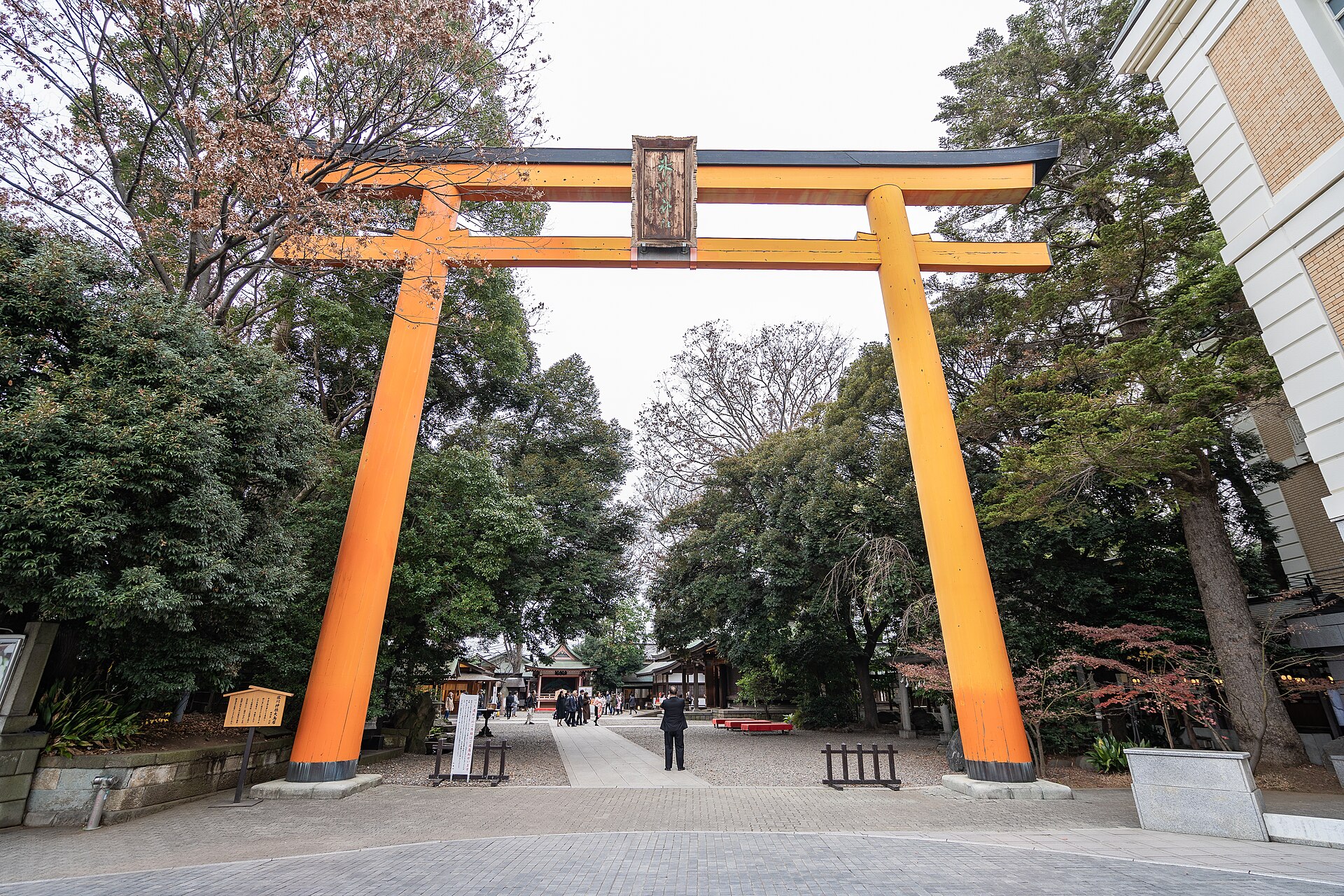 Large orange torii gate standing over a stone path, flanked by tall trees and a white building