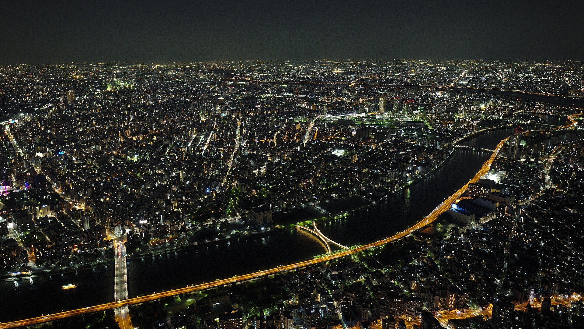 Night aerial view of dense Tokyo city lights, with a river and a glowing bridge winding through