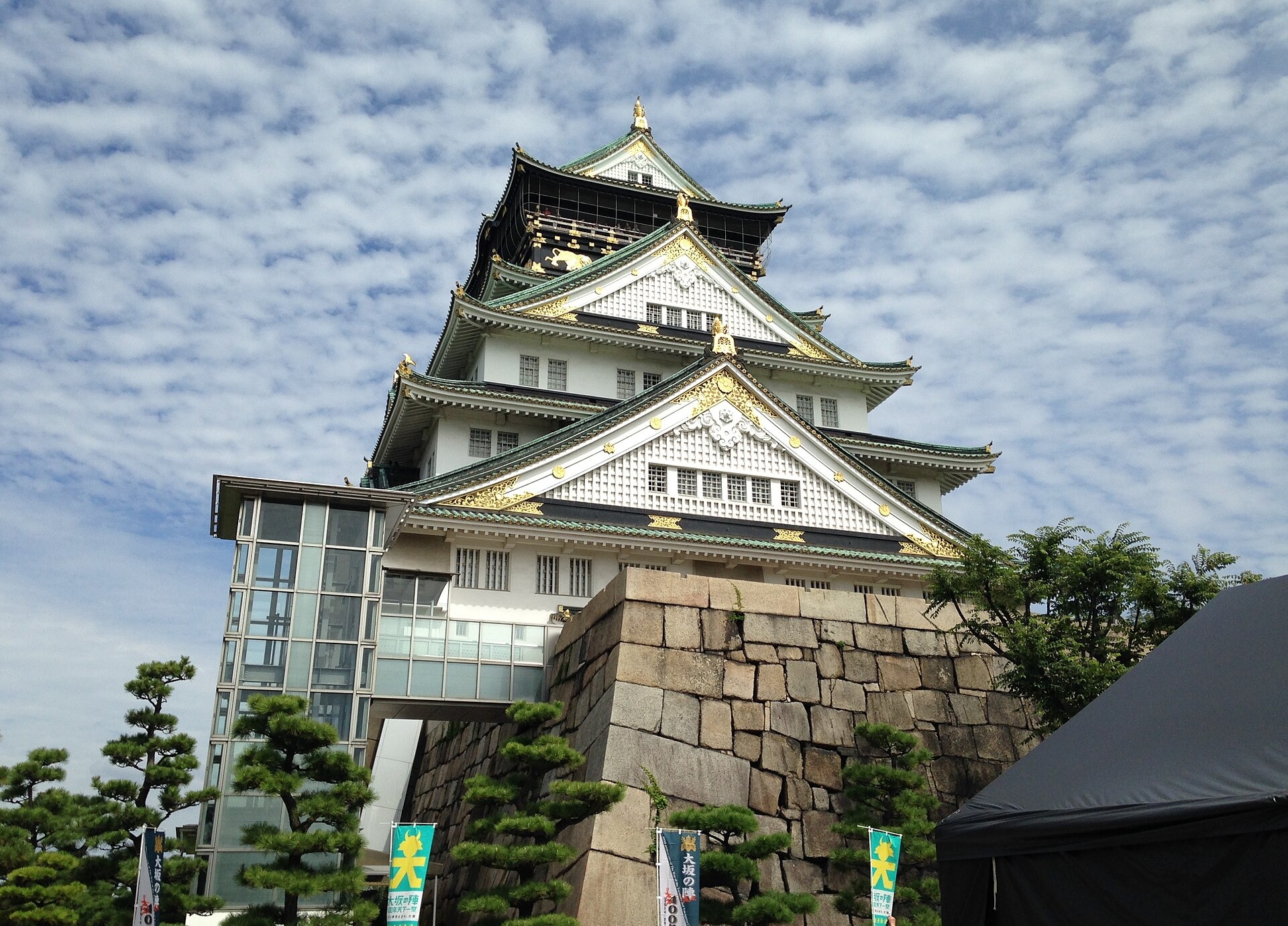 Osaka Castle's white multi-tiered keep with gold ornaments, rising above a stone wall and pine trees under cloudy sky.