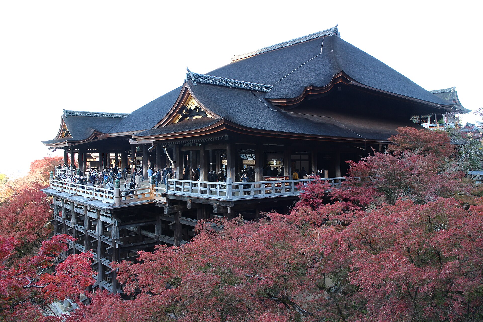 Kiyomizu-dera's wooden stage and dark tiled roof, surrounded by red autumn maple trees, with visitors on the railing