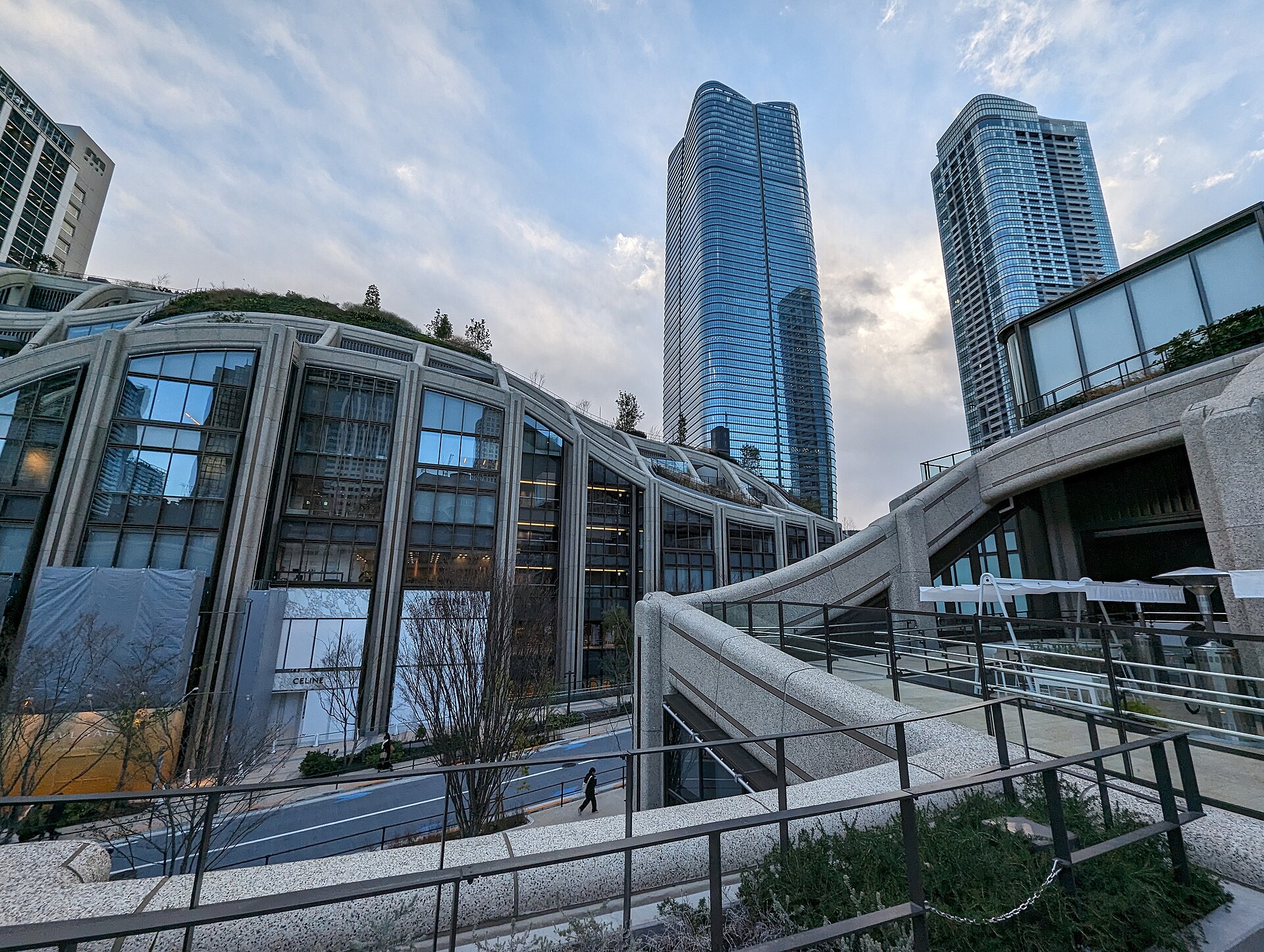 Glass skyscraper and green-roofed curved buildings at Azabudai Hills under a cloudy evening sky