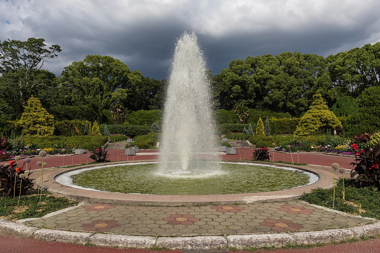 A tall central fountain spraying water in a circular pool, surrounded by green trees, flower beds, and a cloudy sky.