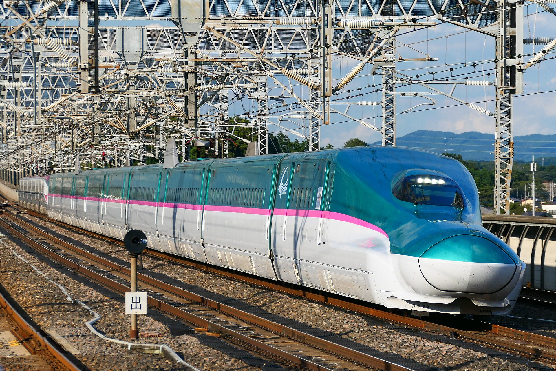 A green, white and pink Shinkansen bullet train speeds along tracks beneath overhead electric wires.