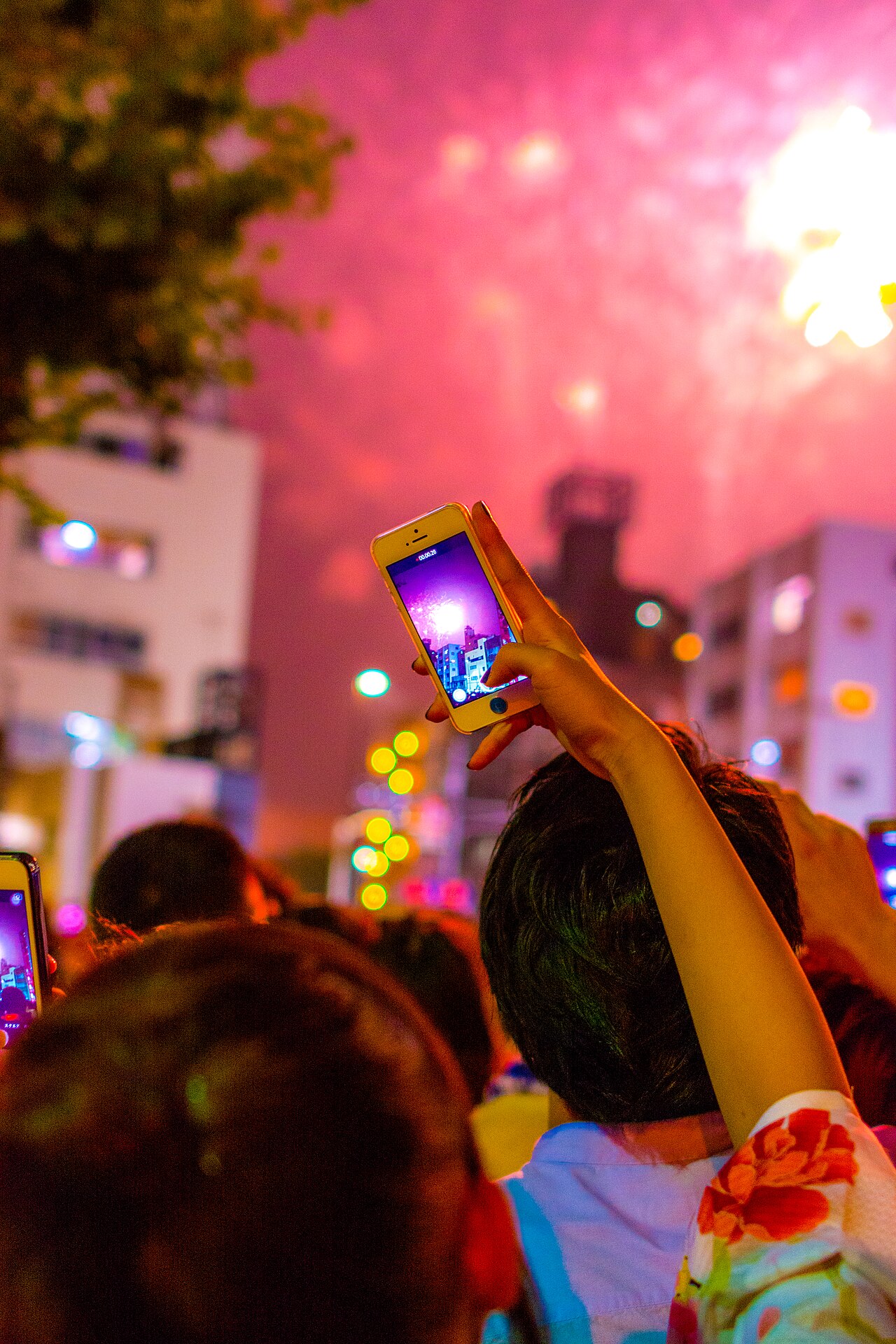 Crowd at night raising phones to film pink fireworks over city buildings