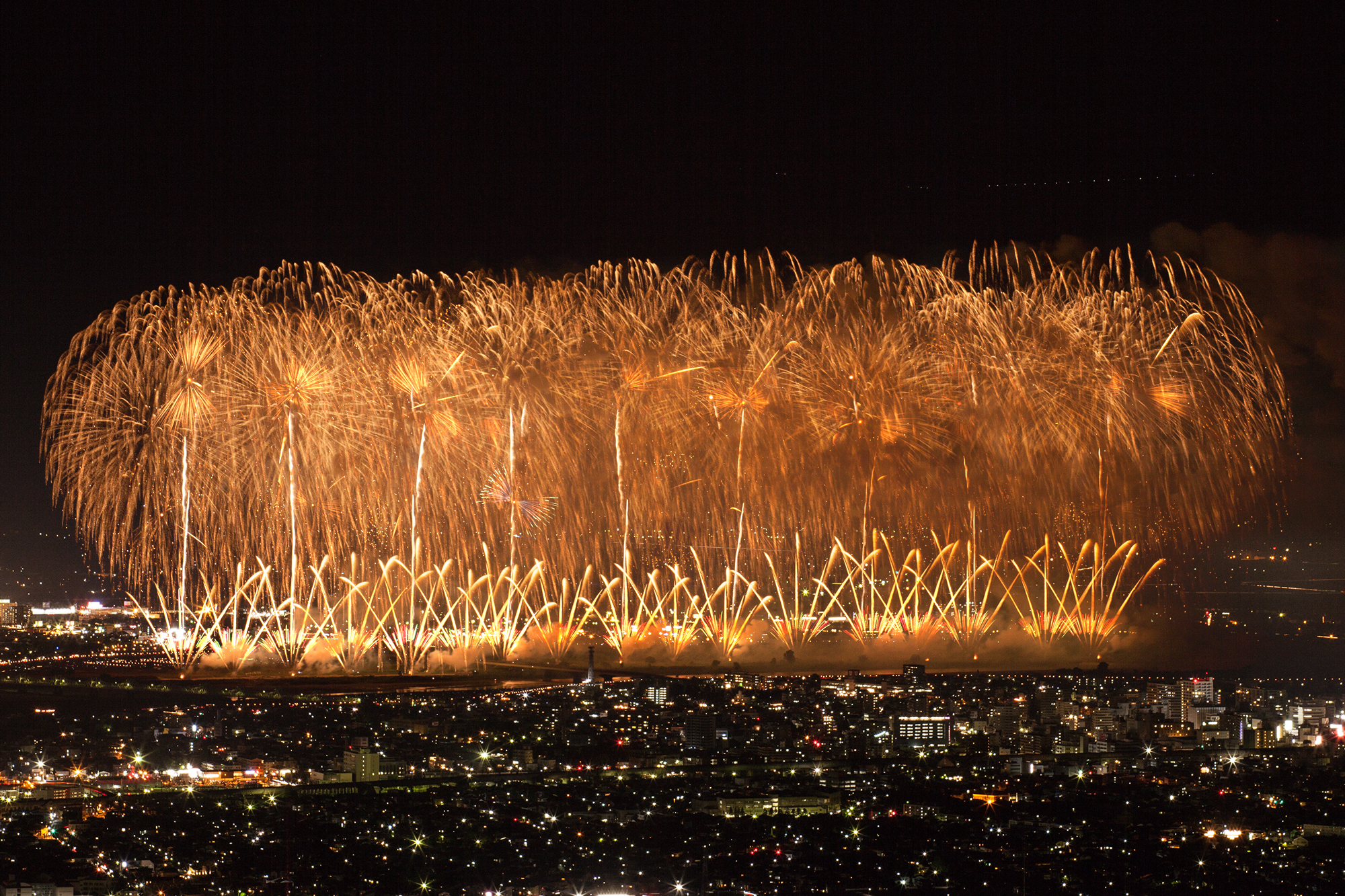 A wide spread of golden fireworks bursting over a night city skyline with lit streets below