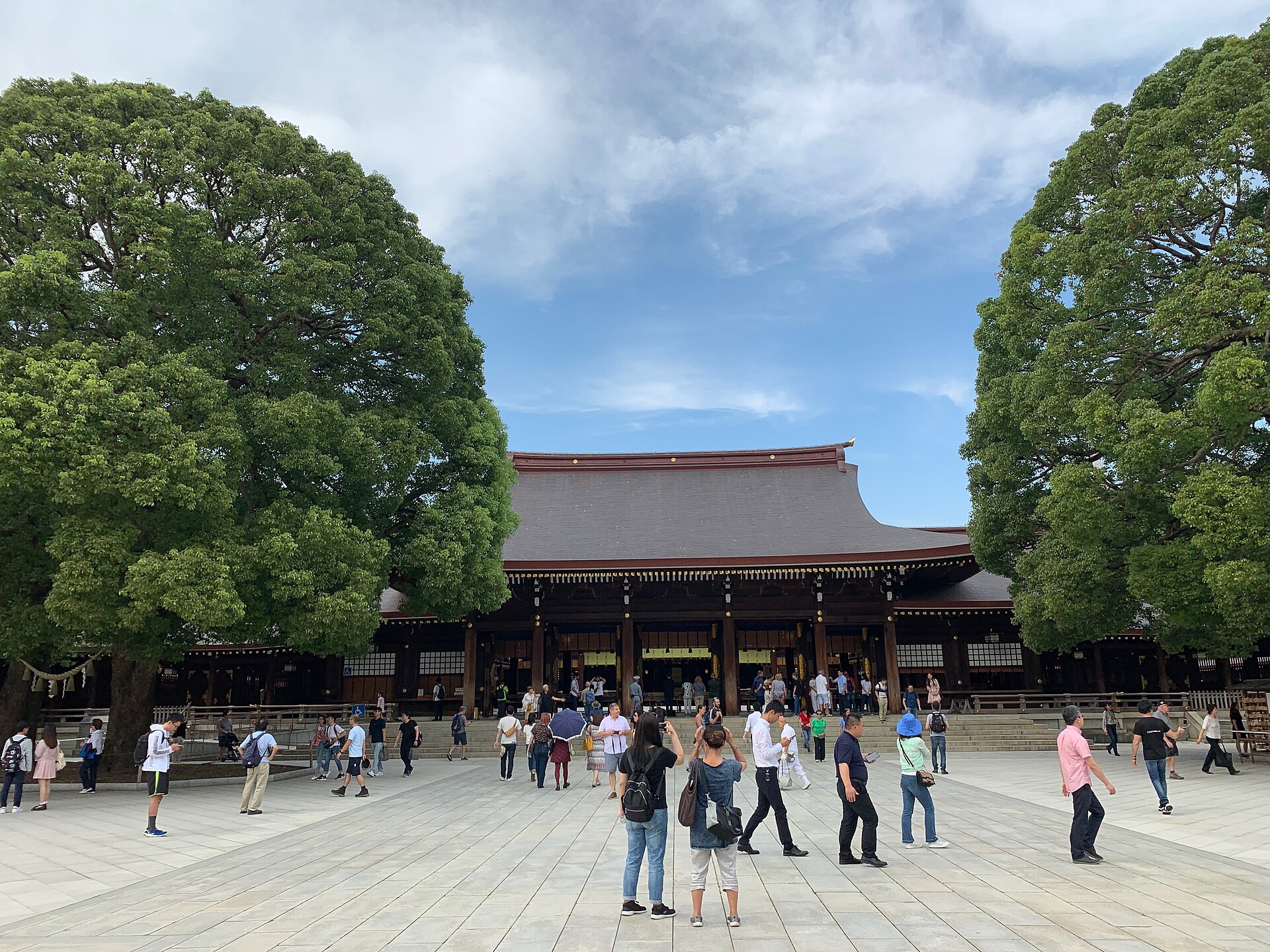 Visitors gather on a stone plaza before the Meiji Shrine main hall, flanked by large trees.