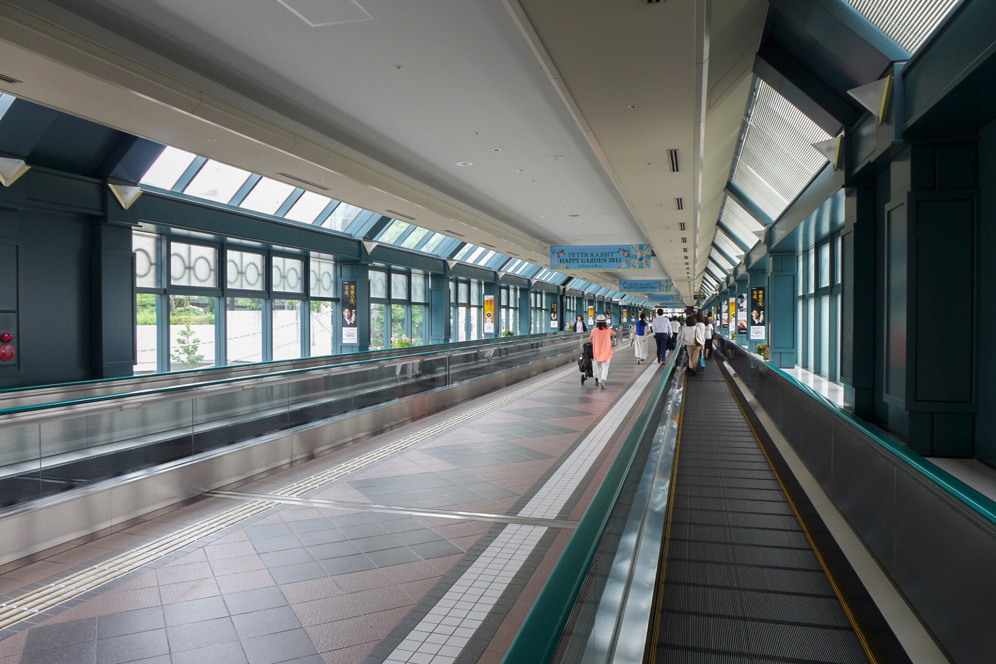 Interior of an elevated covered walkway with moving walkways, glass windows, and pedestrians walking under a skylight