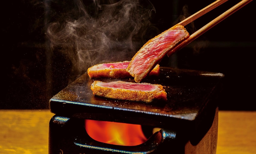 Pink-centered breaded beef cutlet slices on a dark wooden tray over a flame, lifted with chopsticks