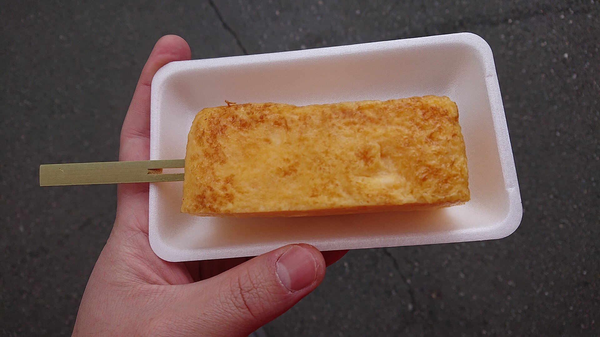 A hand holds a white foam tray with a thick golden rolled omelet on a bamboo skewer, on asphalt