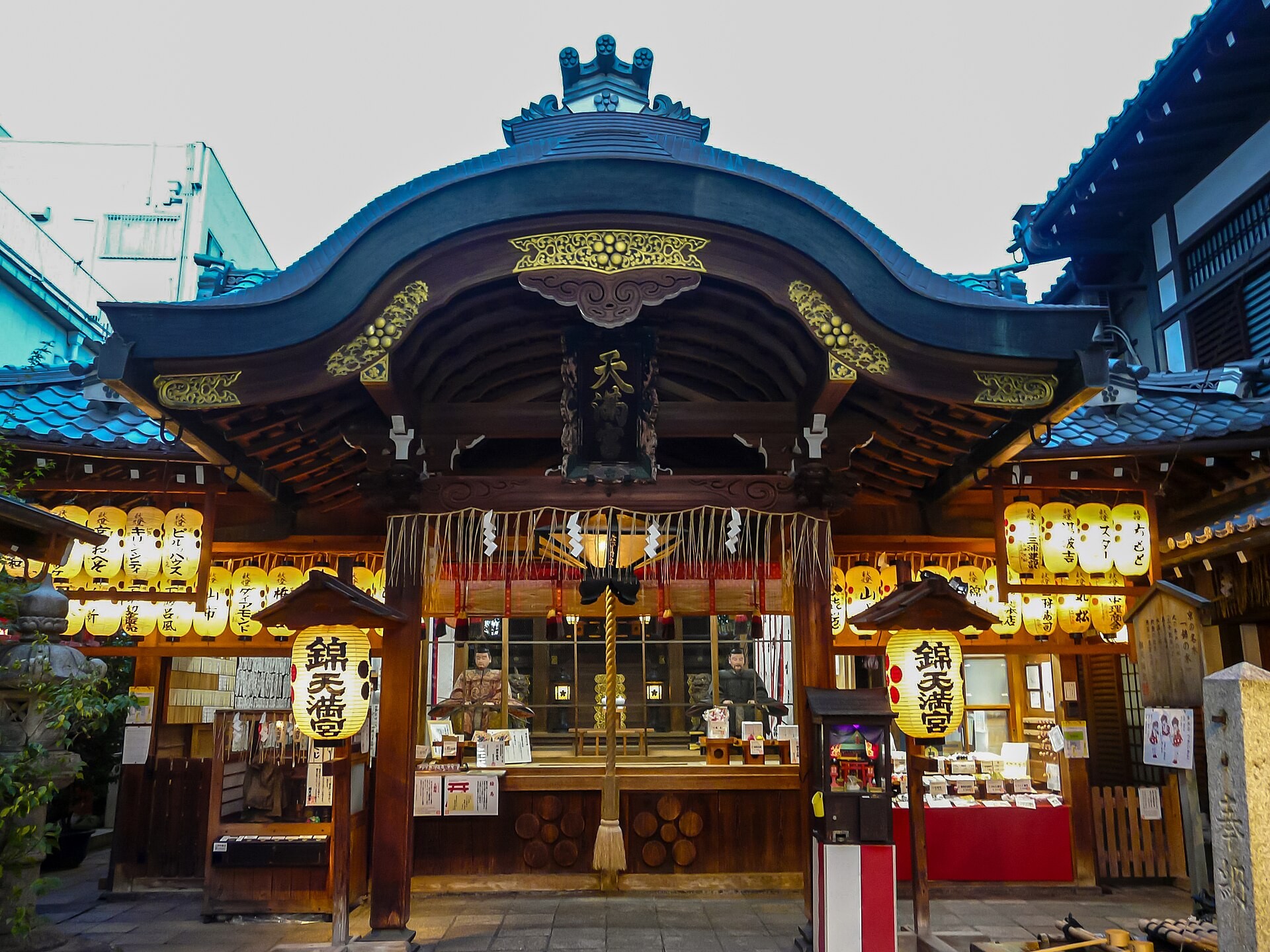 Wooden Nishiki Tenmangu shrine facade with yellow lanterns, white shide streamers, and a blue-tiled roof.