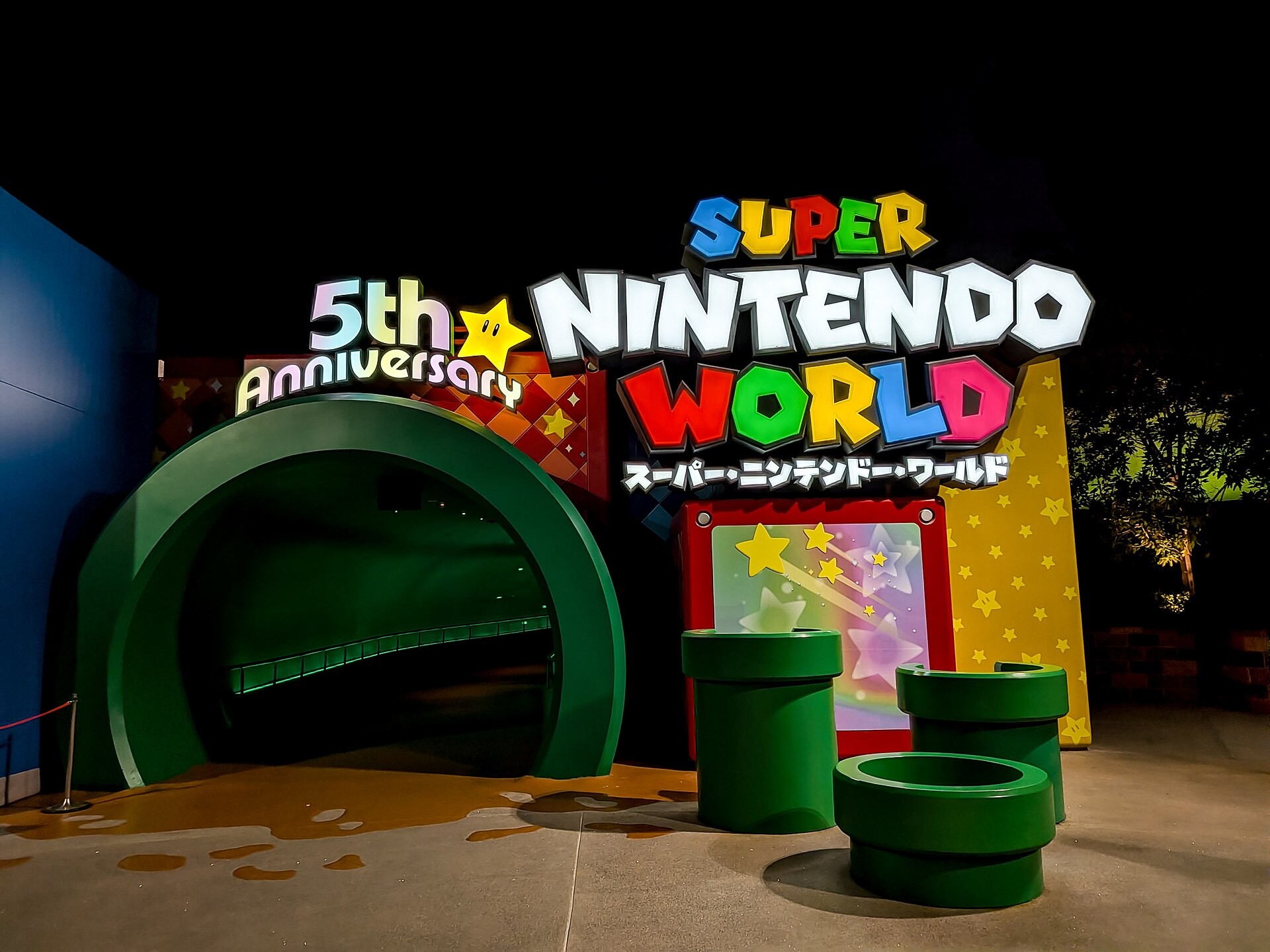 Super Nintendo World entrance at night, with green warp pipes and colorful logo sign