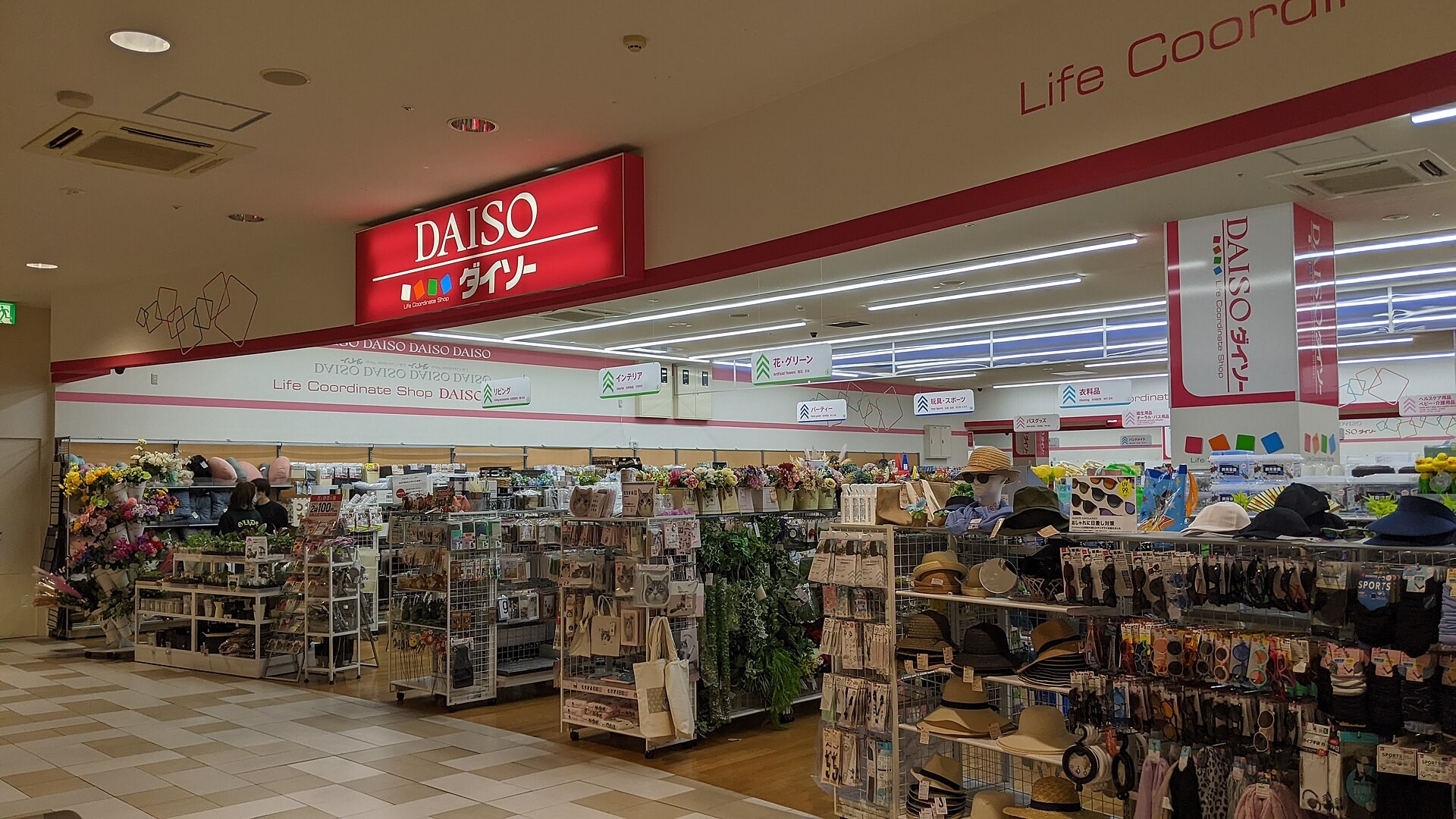 DAISO store entrance with red sign, displaying hats, flowers, and household goods in a mall