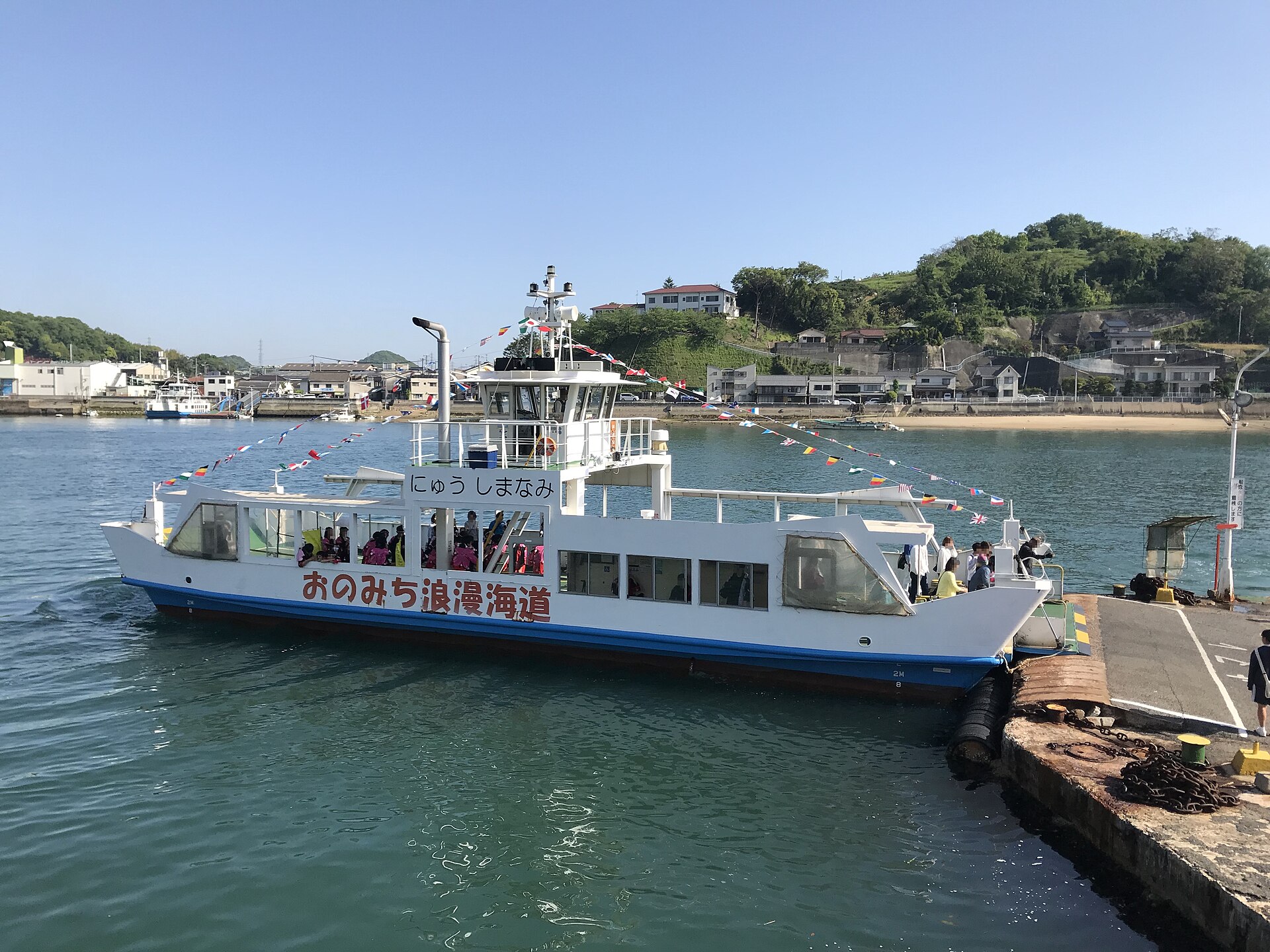 White ferry boat docked at a harbor pier, strung with colorful pennant flags, green hillside town behind