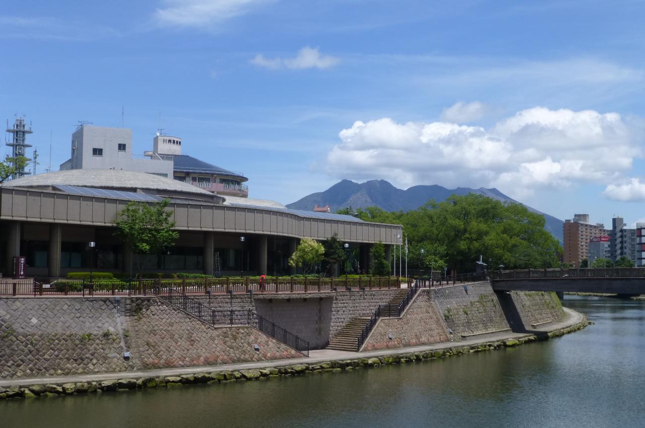 Exterior of the Ishin Furusato Kan museum beside a river, with stone embankments and Sakurajima volcano in the distance under blue sky.