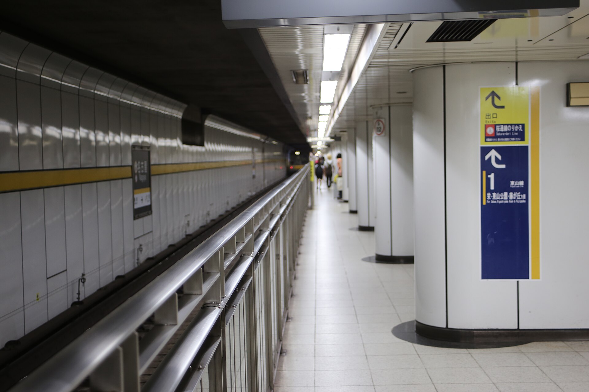 Subway platform with white tiled walls, yellow stripe, and a blue Higashiyama Line direction sign on a pillar.