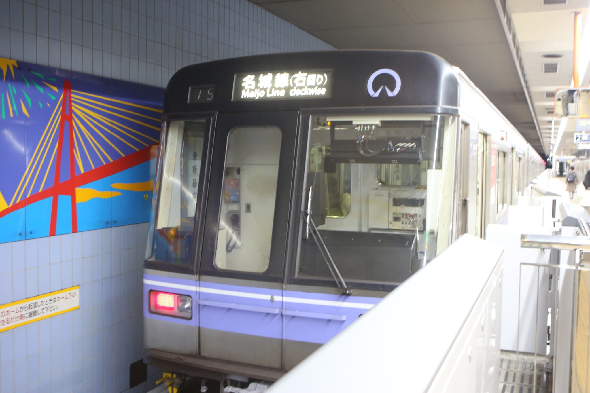 Meijo Line train stopped at a platform beside a blue bridge mural