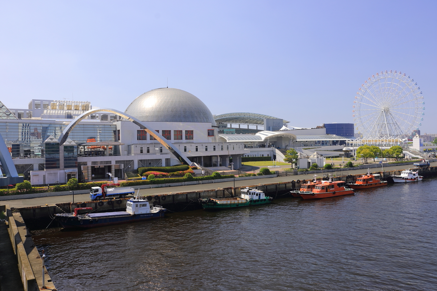 Harbor waterfront with moored boats, a domed building, and a white Ferris wheel under clear sky