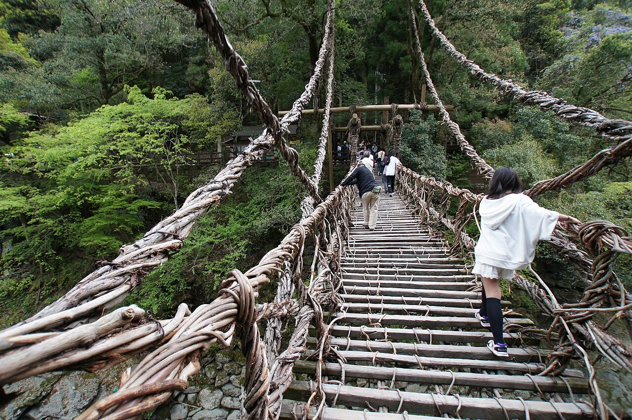 Visitors walk across a vine-woven suspension bridge with gaps between wooden planks, surrounded by green forest.